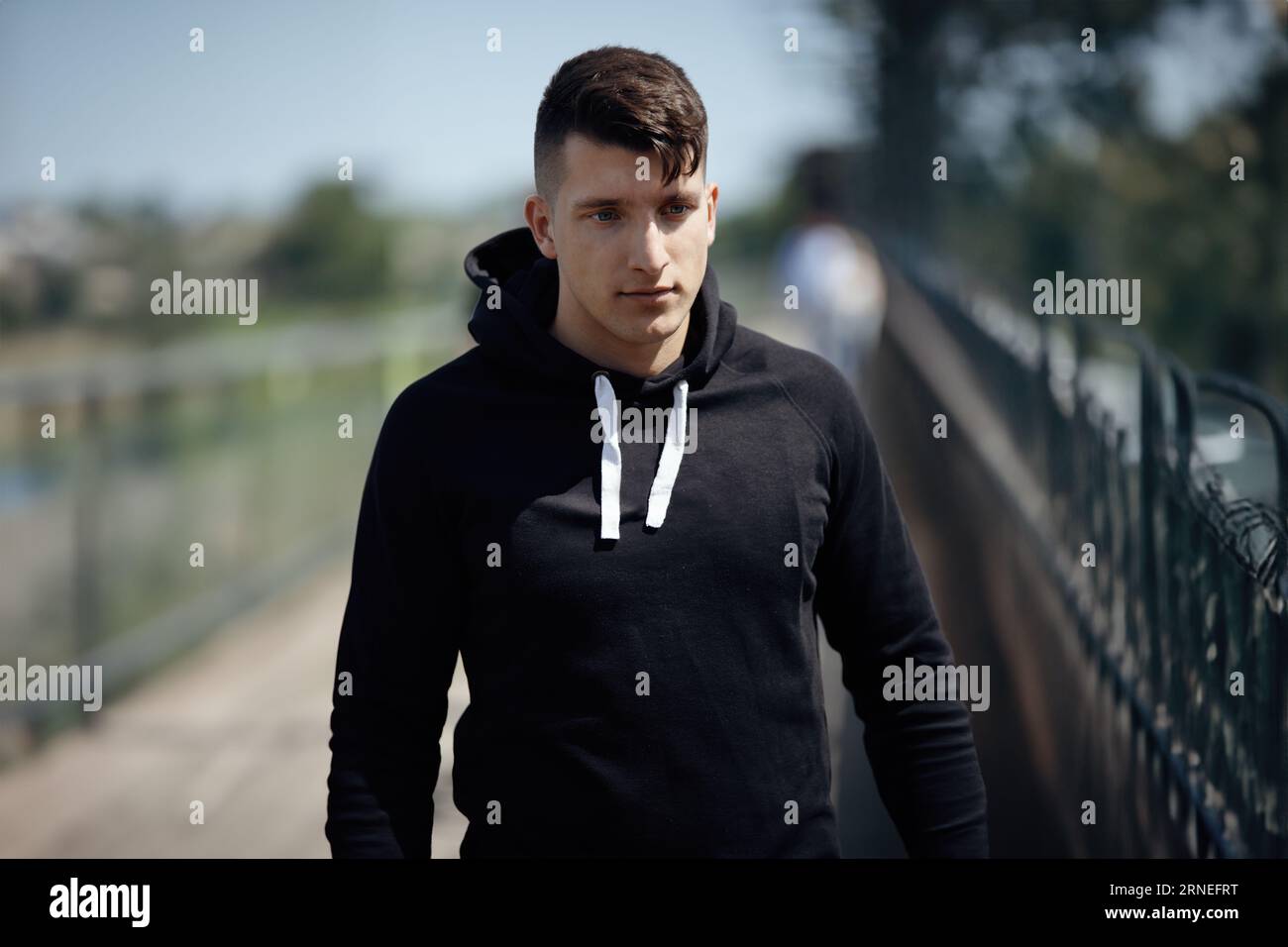 Young Man with black sweater walk outside in park in midday sun and look down to side with a neutral and serious face expression Stock Photo