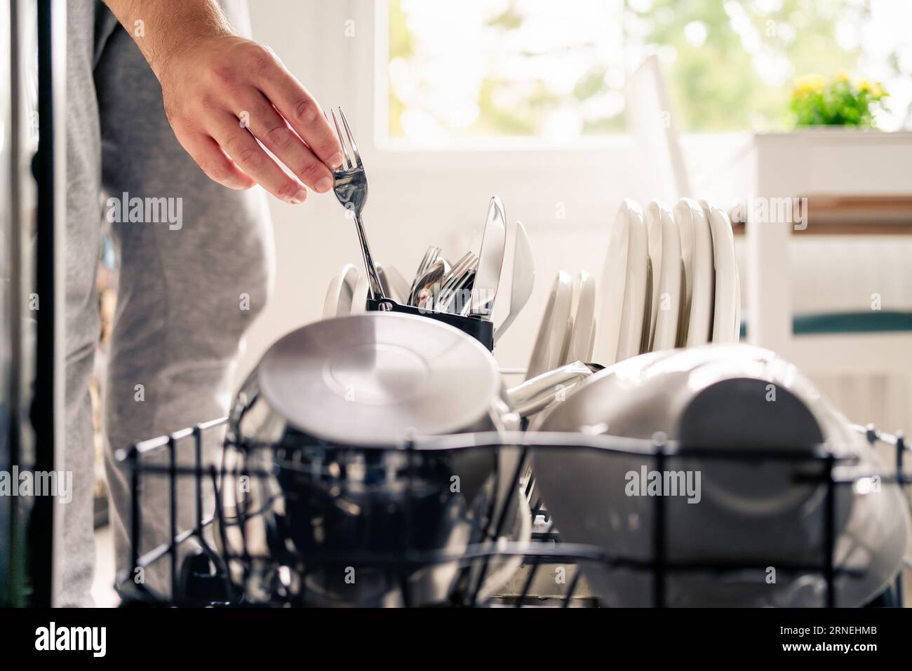 Dish washer machine in kitchen. Man loading dishwasher. Washing plates. Fork in hand. Full of cutlery. Clean or dirty. Pot and tableware. Stock Photo