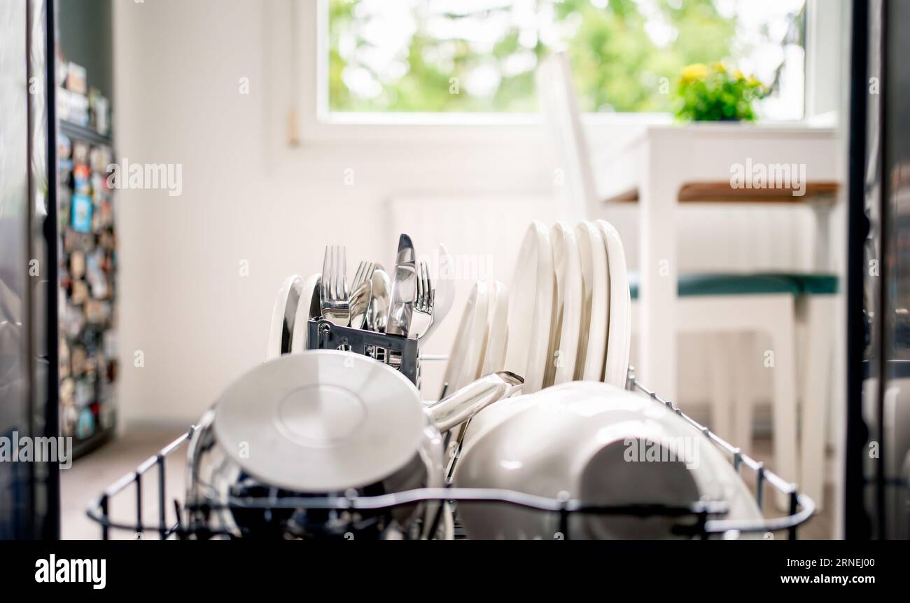 Dish washing machine. Inside an open dishwasher, view to kitchen. Clean plates, utensils, cutlery and pot in washer rack and shelf. Basket with fork. Stock Photo