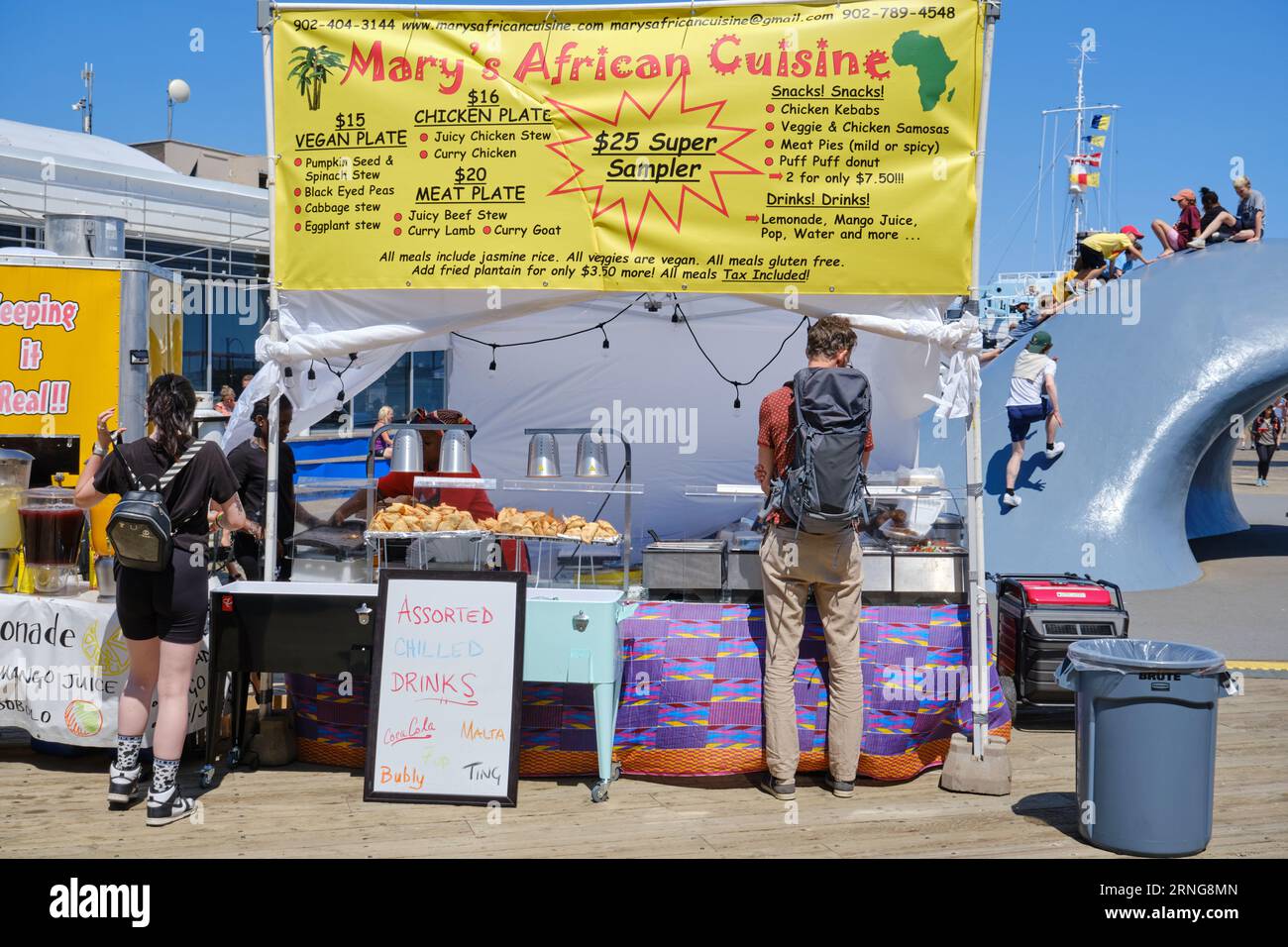 Mary's African Cuisine food stand on the Halifax waterfront during festival, with kids climbing wave in background Stock Photo