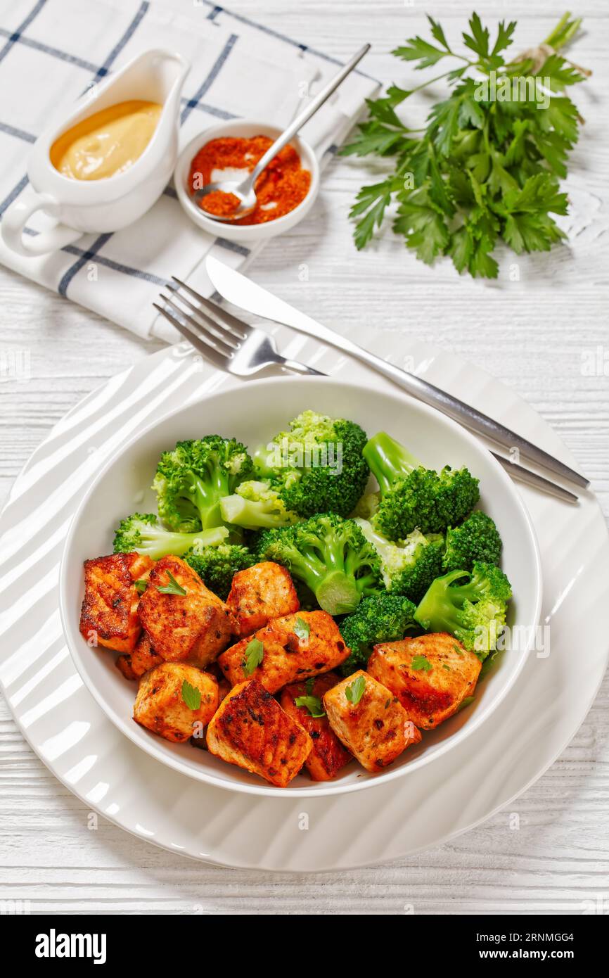 broiled salmon chunks with steamed broccoli florets in white bowl on white wood table with sriracha mayo sauce, vertical view from above Stock Photo