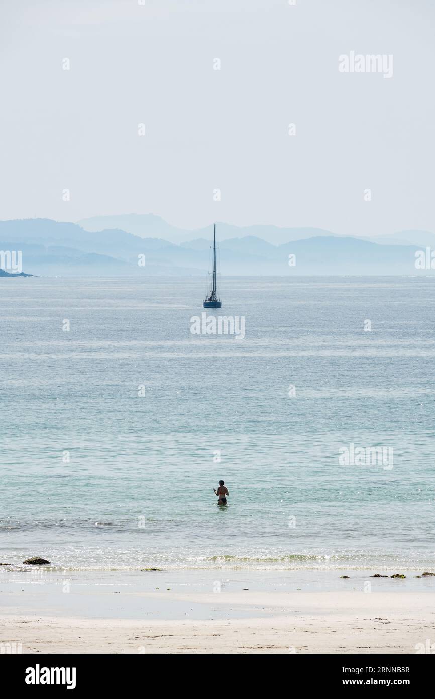 PONTEVEDRA, SPAIN - APRIL 8, 2023: A lonely middle-aged woman on a beach in the Ria de Pontevedra in Galicia on a foggy spring day, with a blue sailbo Stock Photo