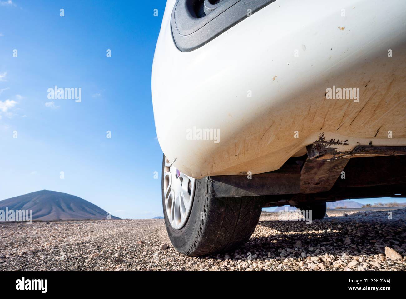 A white car parked on a gravel road in the desert. The car is alone on the road, and the only other thing in sight is a small bush. The sky is blue, a Stock Photo