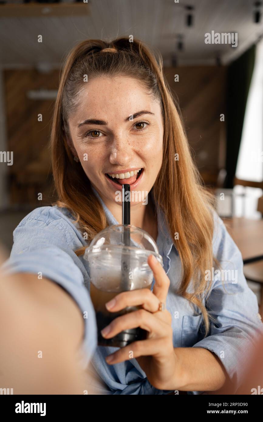 Young freckled woman drinking bubble tea and taking a selfie in a cafe