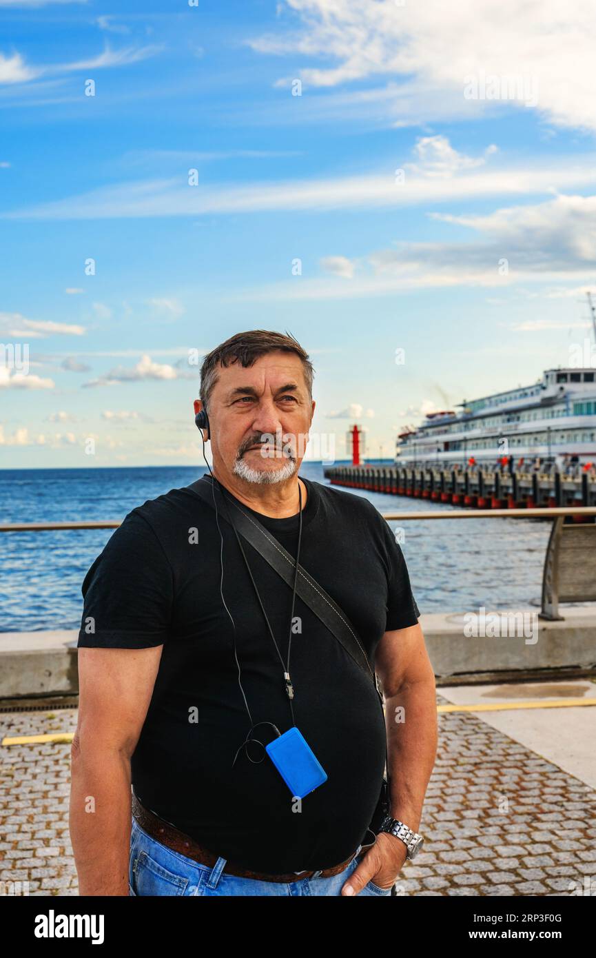 A man in his 70s with a tourist earpiece and an eradio guide standing on a pier in the background of a river cruise liner. Travelling pensioners. Stock Photo