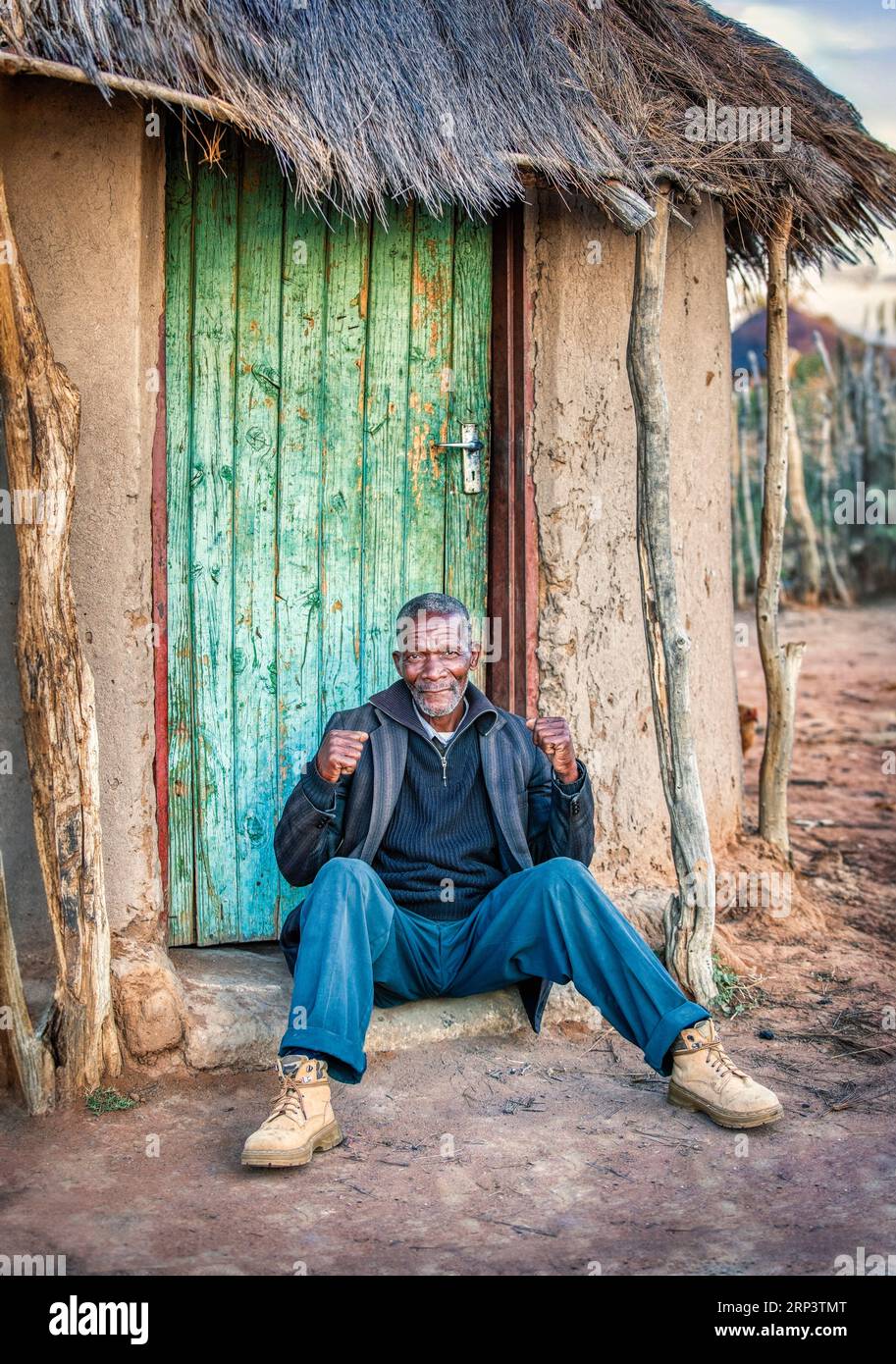 ld african villager portrait sited on the doorstep , gesturing victory with his fists, outdoors in the yard of his home Stock Photo