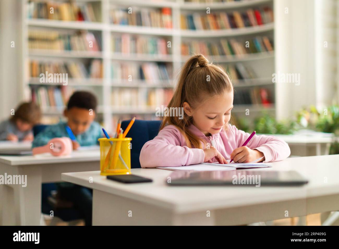 Group of smart school children sitting at desks in classroom or library at primary school, writing in their workbookk Stock Photo