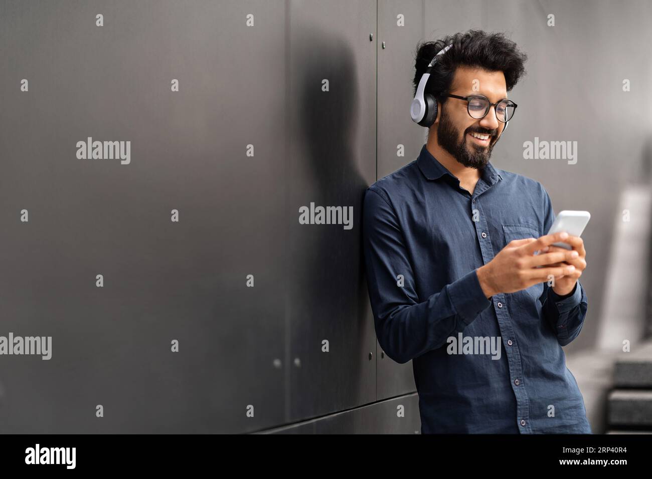 Millennial eastern man standing by black wall, using phone, headphones Stock Photo