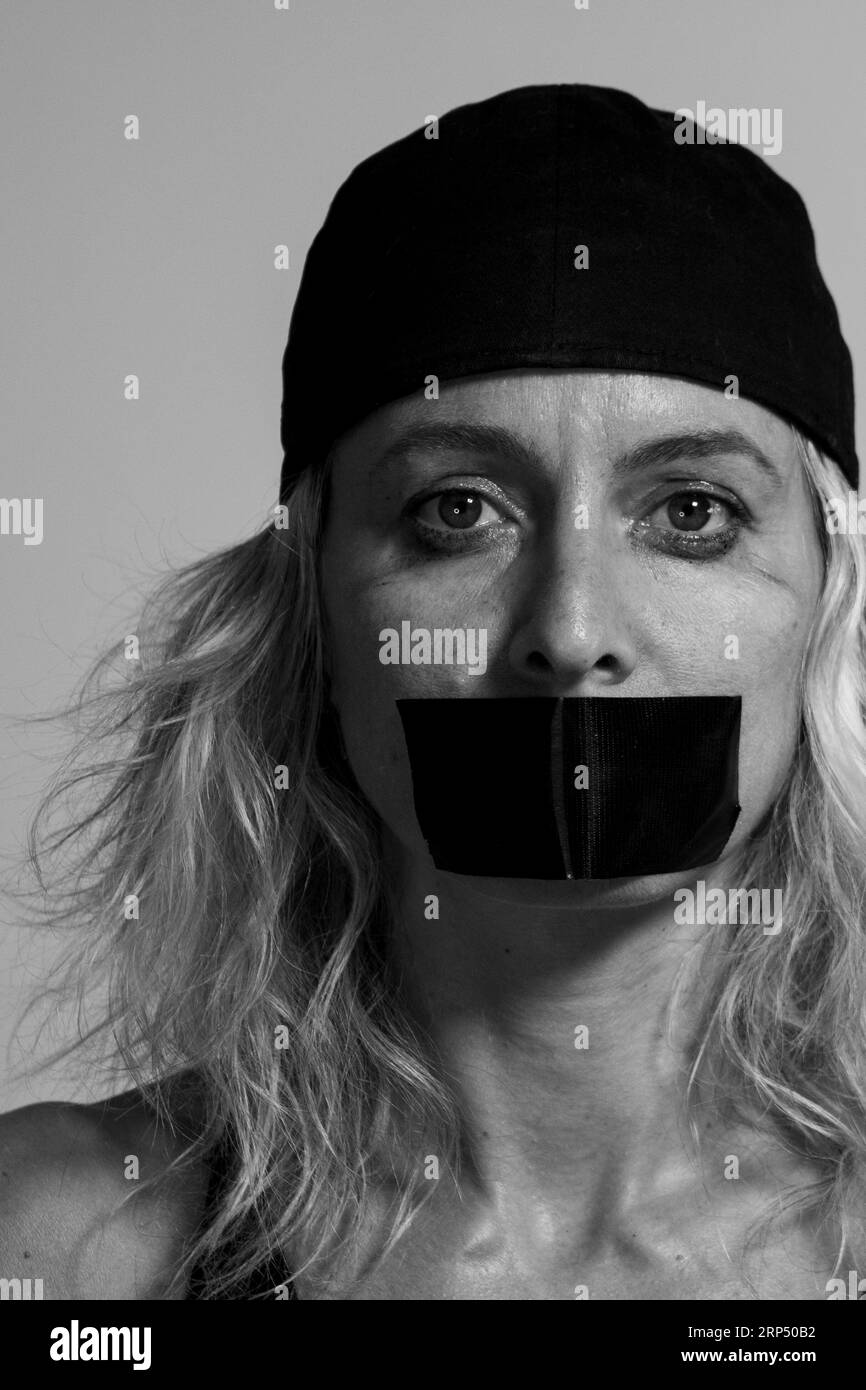 Light-eyed blonde woman with a cap and her mouth taped so she can't speak Stock Photo