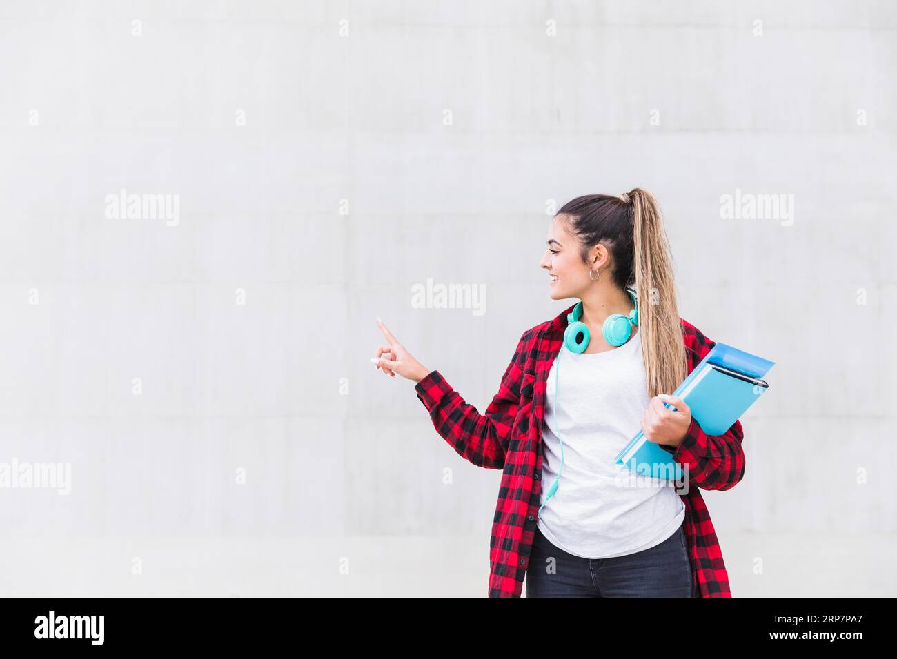 Portrait smiling female university student holding books hand pointing her finger white wall with copy space Stock Photo