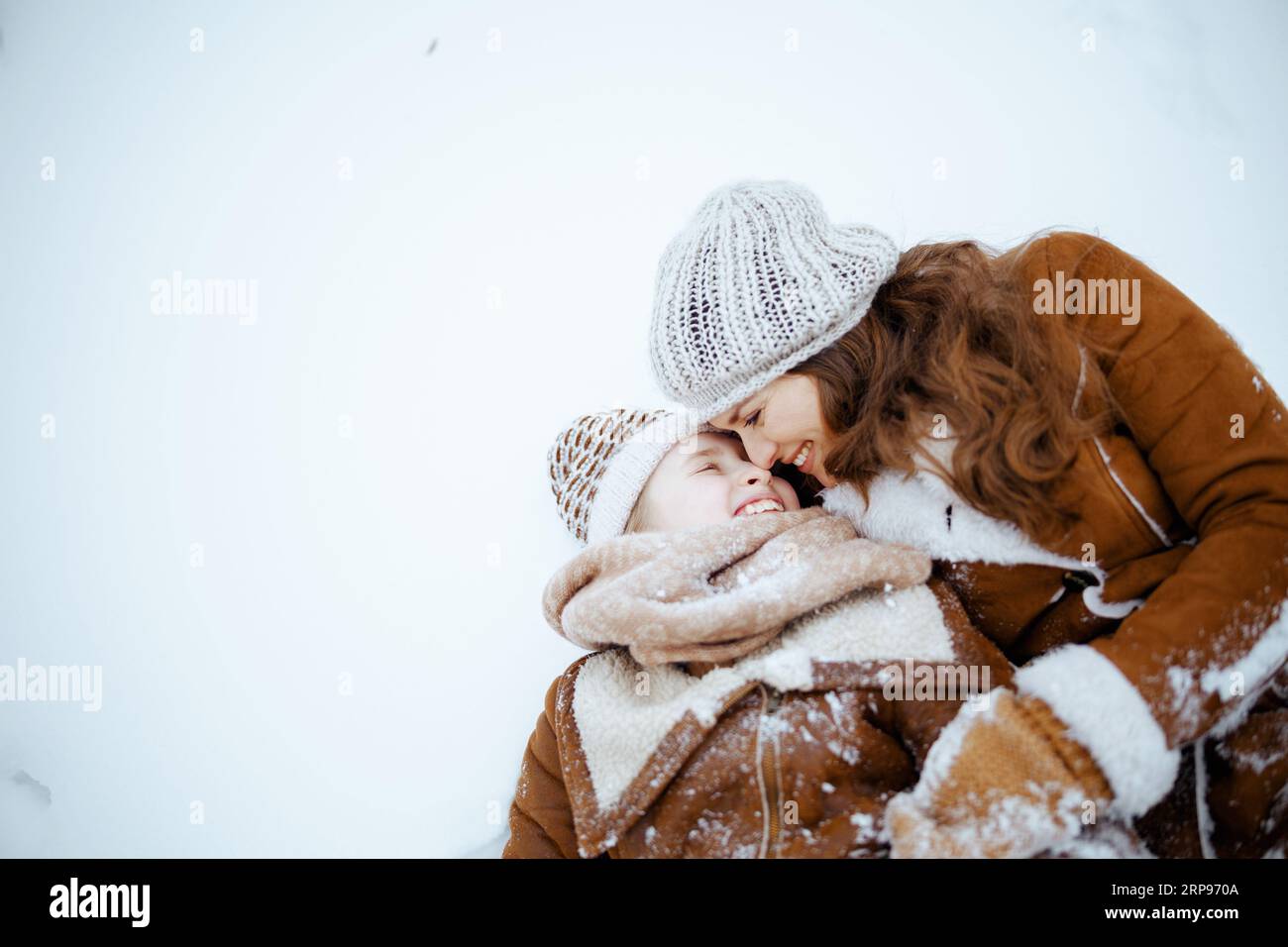 happy elegant mother and daughter in coat, hat, scarf and mittens laying in snow outdoors in the city park in winter. Stock Photo