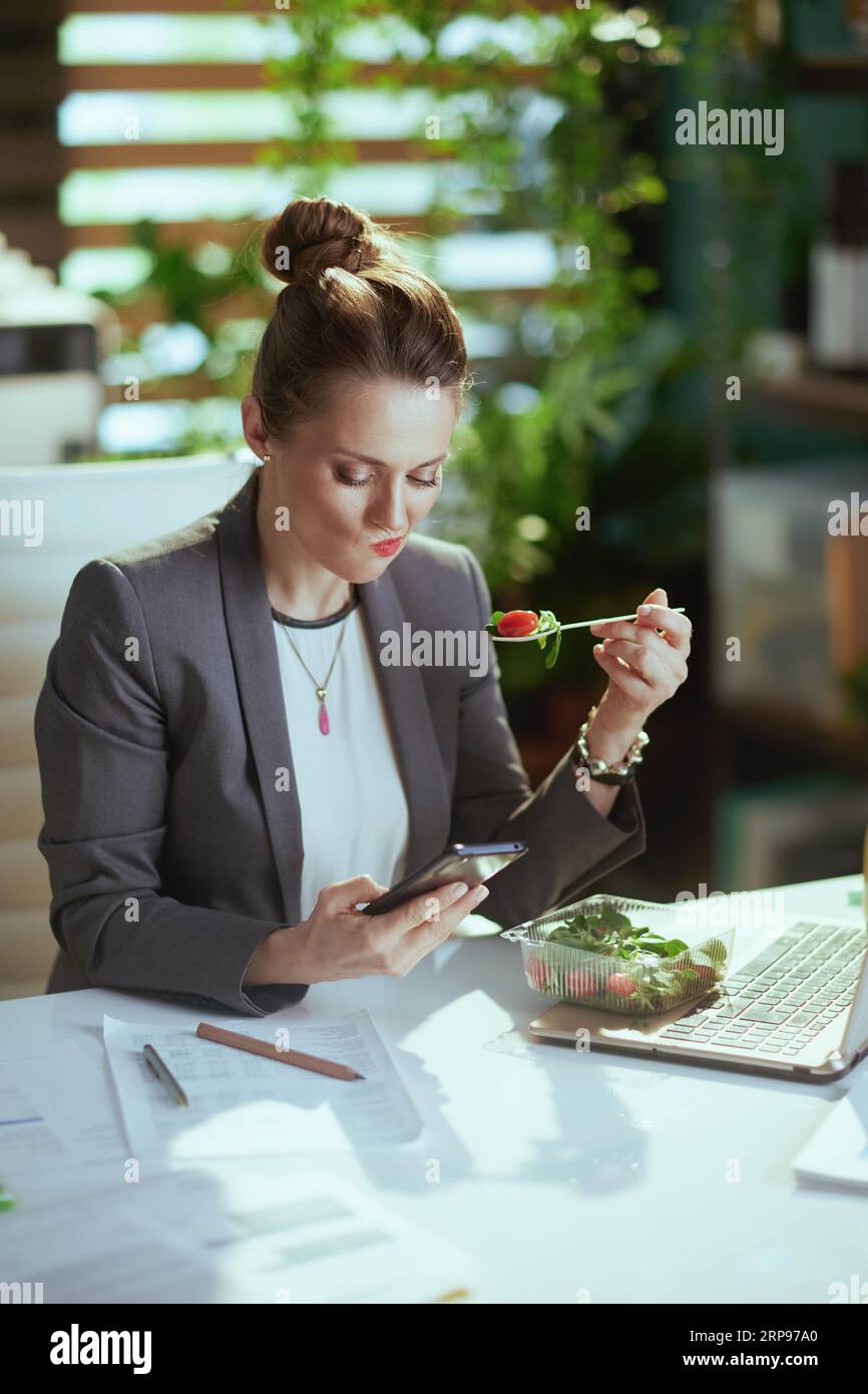 Sustainable workplace. sad modern 40 years old woman worker in a grey business suit in modern green office with laptop eating salad and using smartpho Stock Photo