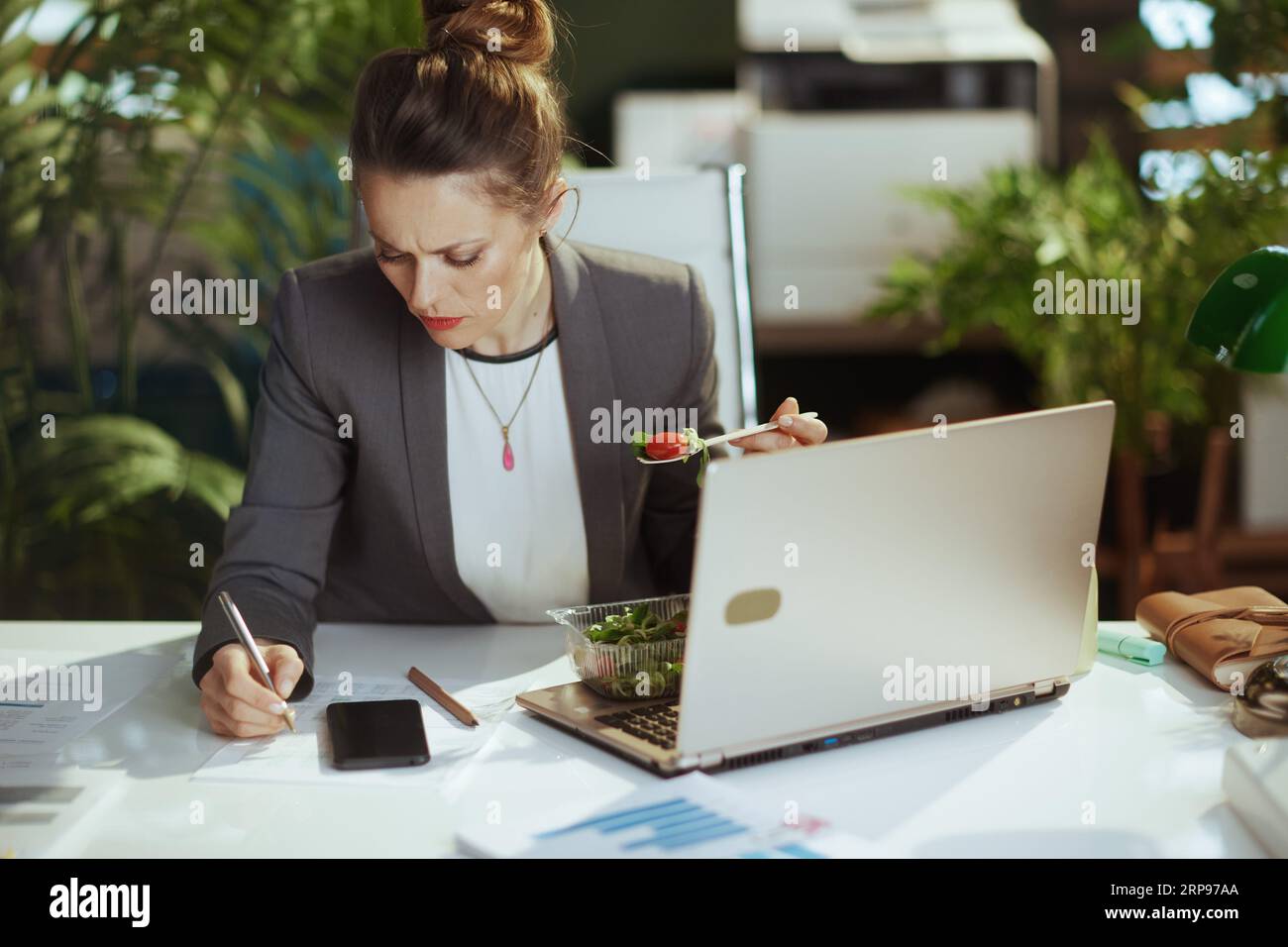 Sustainable workplace. concerned modern business woman in a grey business suit in modern green office with laptop eating salad and using smartphone. Stock Photo