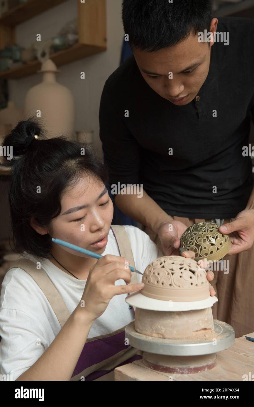 (190520) -- LONGQUAN, May 20, 2019 (Xinhua) -- Lin Song (R) and his wife Ji Jing discuss the making of celadon incense burner caps at his studio in Longquan, east China s Zhejiang Province, May 20, 2019. Born in 1993, Lin Song is a native of Longquan, home to China s best celadon wares. He received professional training in both his hometown and Jingdezhen, another important porcelain hub of China. Lin had also followed other celadon artisans for years before setting up a studio back home in 2014, focusing on the replication of classical celadon incense burners. Lin s personal collection has gr Stock Photo