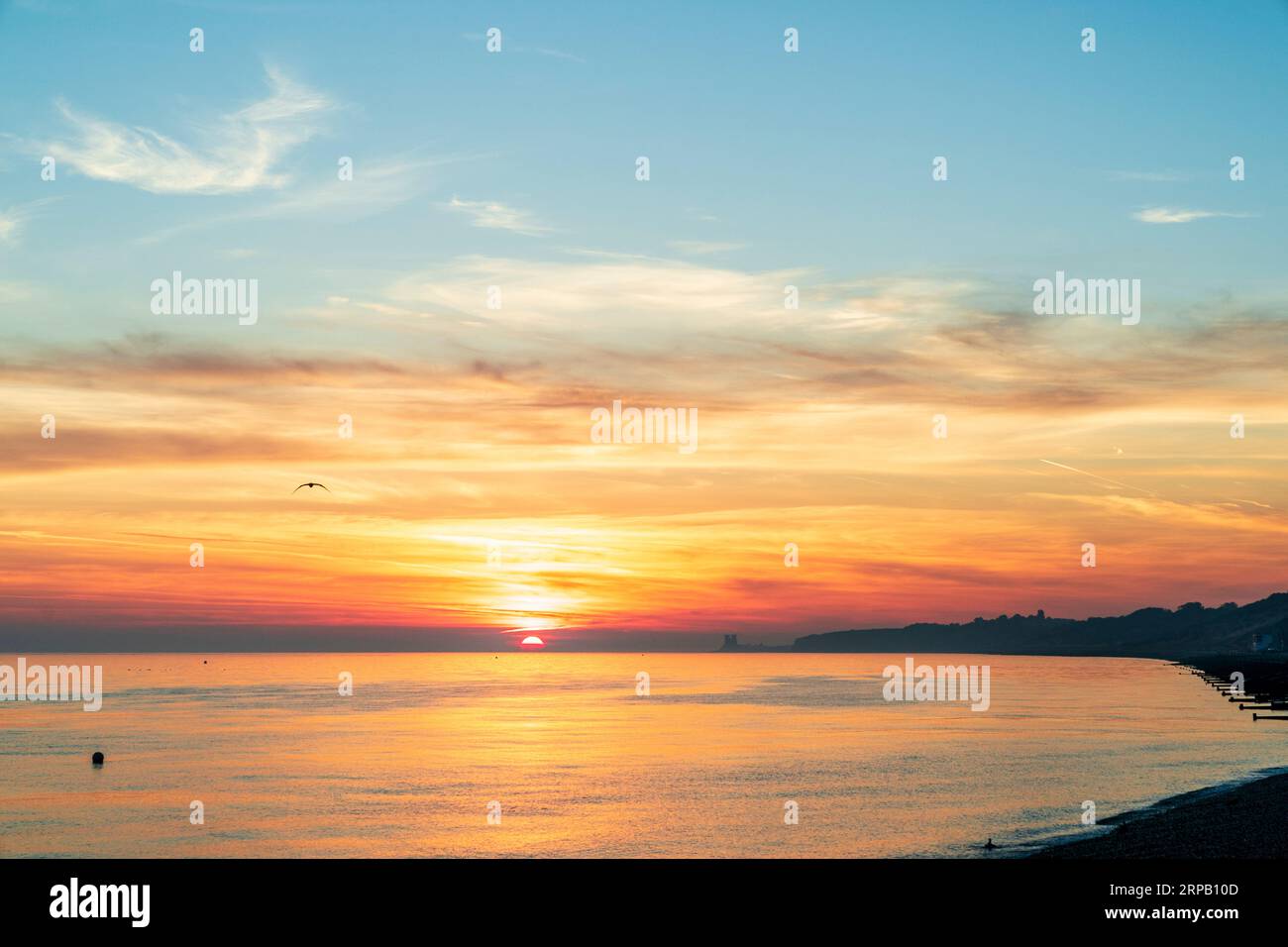 The sunrise seen from the beach at the seafront at Herne Bay on the north English Kent Coast. A blue sky with a vivid layer of red and orange clouds near the horizon, the orange being reflected in the very calm sea. the sun half risen over the sea. Stock Photo