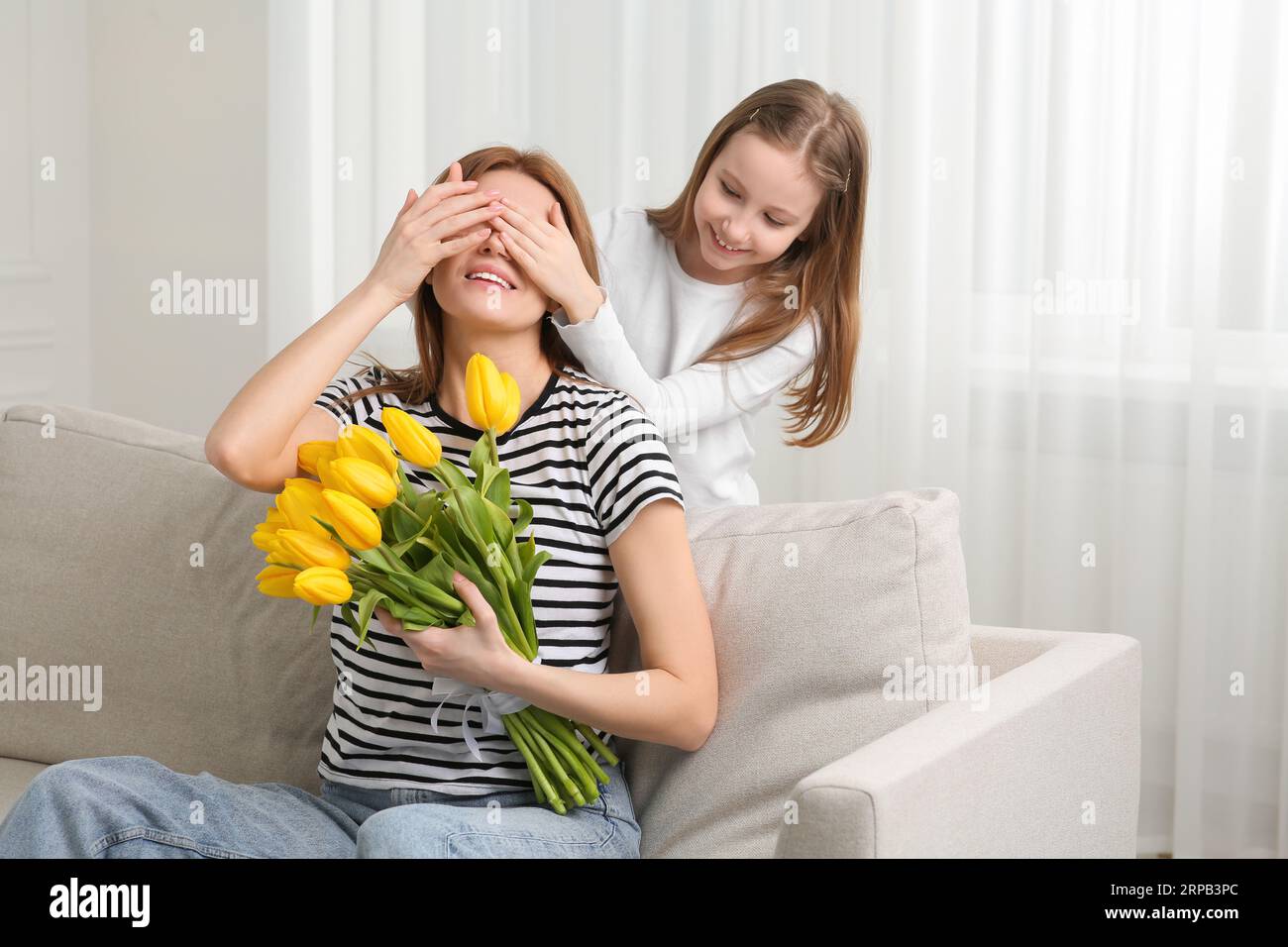 Smiling young adult daughter makes an unexpected surprise to her middle  aged attractive mom, gives her a gift, covering her eyes with self hand,  sitting at living room. Friendship of mom and