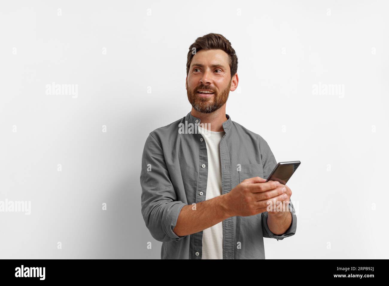 Smiling man looking at side and holding mobile phone while standing on white background Stock Photo