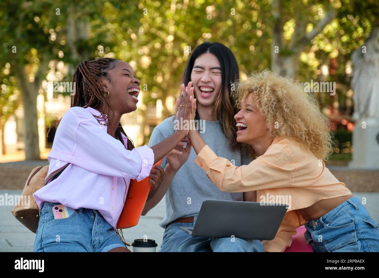 Three happy multiracial friends high five and laughing because got a perfect test score at a park. Stock Photo
