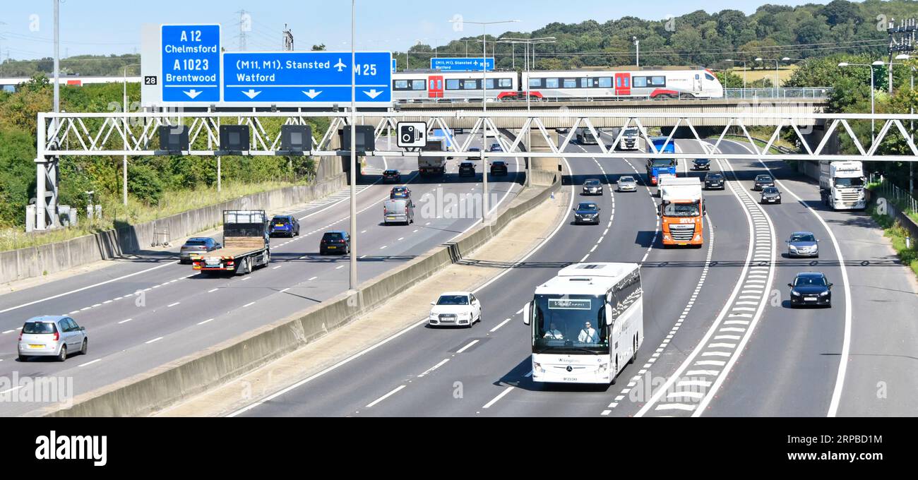 M25 motorway gantry signs railway bridge & Greater Anglia passenger train crossing above road traffic junction 28 for A12 Brentwood Essex England UK Stock Photo