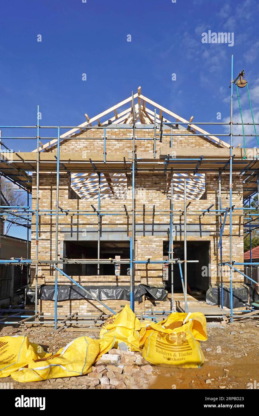 Front elevation new build brick detached house on infill plot takes shape gable wall & roof under construction wrapped in scaffolding Essex England UK Stock Photo