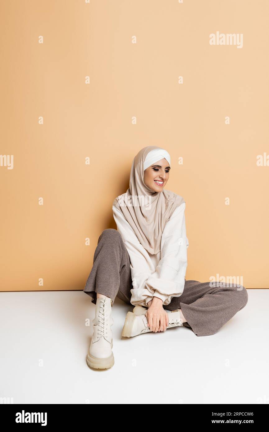 carefree muslim woman in fashionable casual outfit and hijab sitting on beige, full length Stock Photo