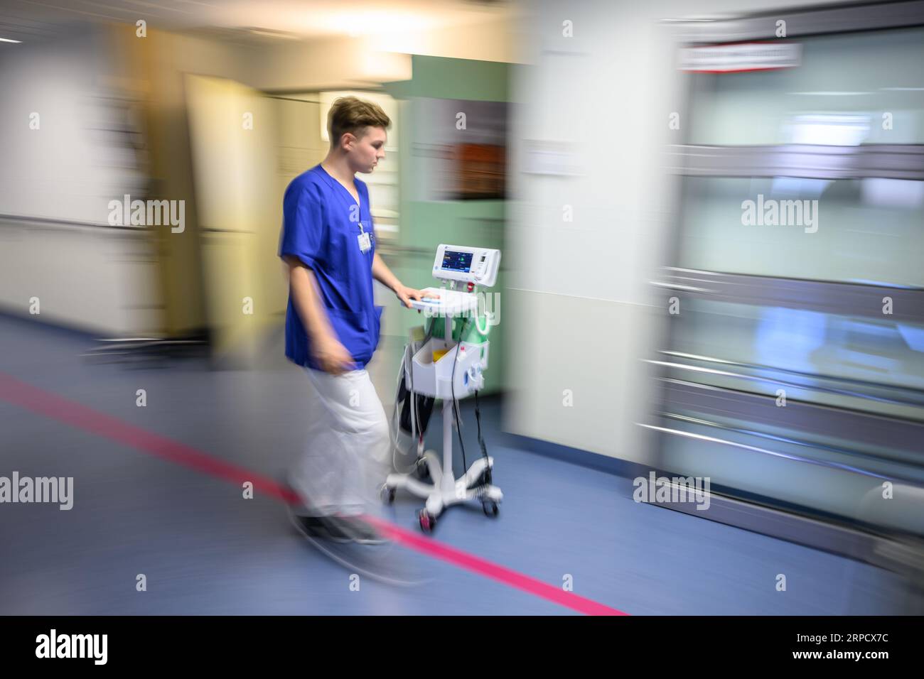 04 September 2023, Saxony, Dresden: Nursing specialist Florian von Rechenberg, one of over 90 trainees who have just passed their exams, pushes a blood pressure monitor across the corridor in the acute geriatrics ward at Dresden University Hospital (long exposure shot). 'One hospital - 17 training professions' - This is the motto under which the University Hospital Carl Gustav Carus Dresden is launching the application phase for training positions in the coming year. Almost 200 positions are available in medical, nursing, therapeutic and administrative areas. Photo: Robert Michael/dpa Stock Photo
