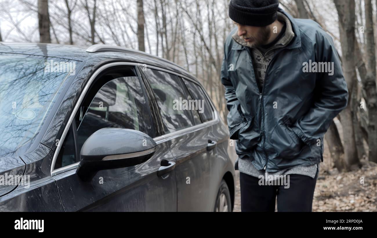 Male tramp bum looks through the slightly open window of auto's door and walks away. Vehicle with alarm system. Beggar. Property insurance against the Stock Photo