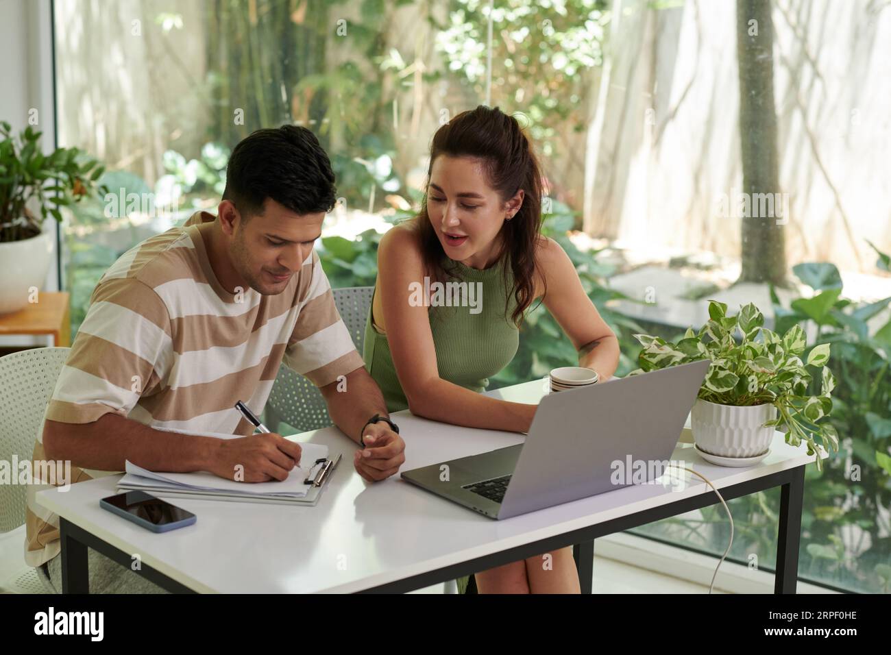 Two young intercultural colleagues organizing work or analyzing online data while man making notes on paperwork during discussion Stock Photo