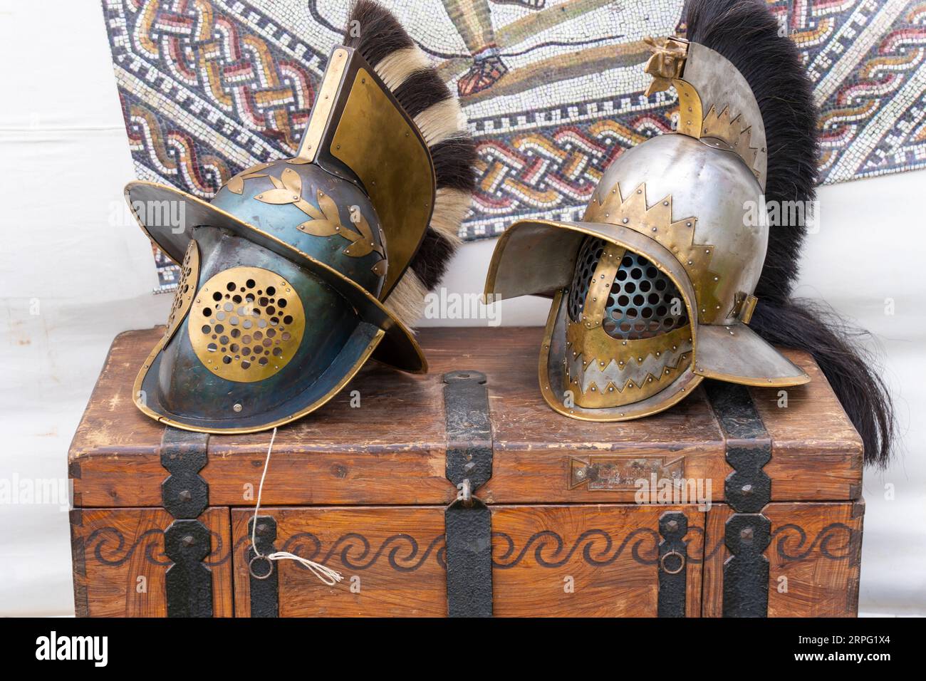 2 ancient roman gladiator helmets on a brown wooden chest with a mosaic image in the background Stock Photo