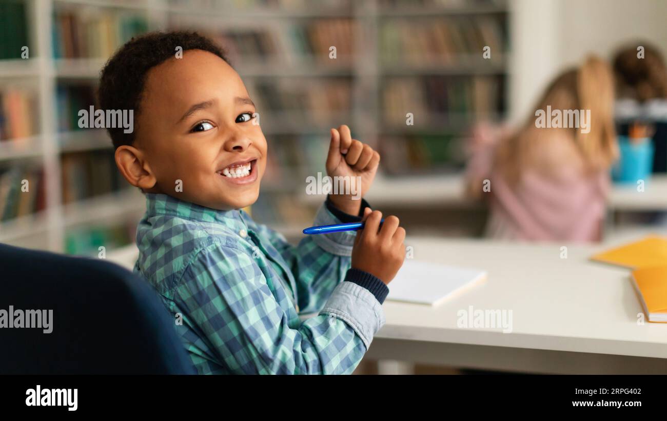 Positive black primary schoolboy sitting at desk in classroom, looking and smiling at camera, panorama, free space Stock Photo