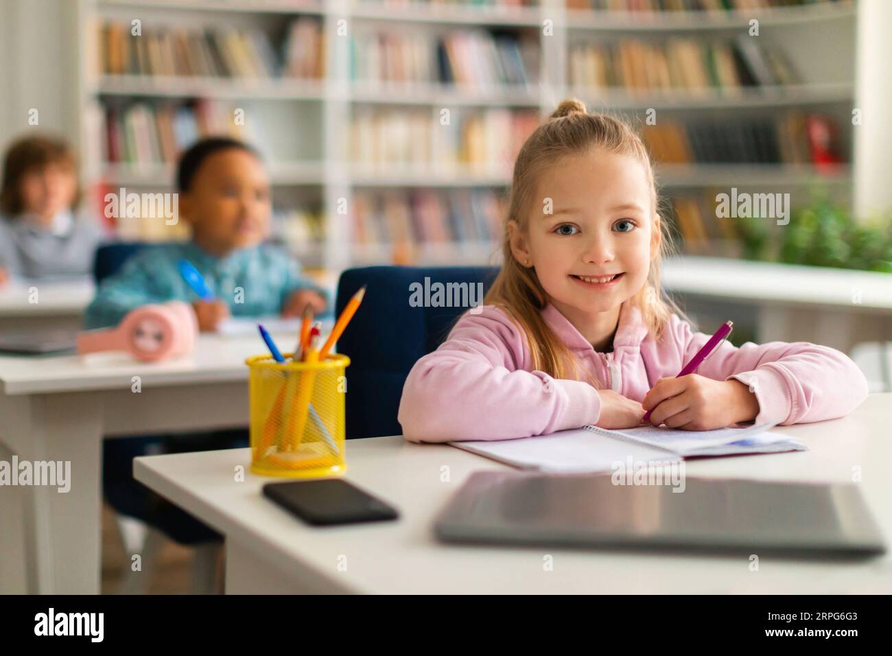 Happy european school girl smiling at camera, sitting at desk in classroom with classmates on background Stock Photo
