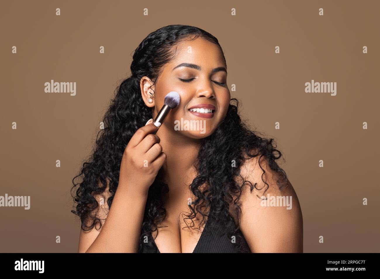 Young happy brazilian plus size woman doing contouring apply blush or highlighter on cheeks with makeup brush Stock Photo