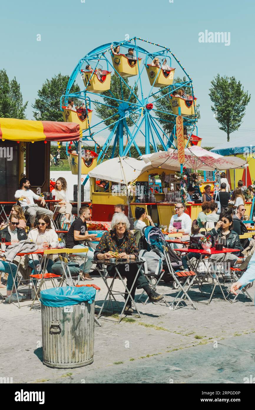 People enjoy food sitting at tables near food stalls at Rollende Keukens food festival, with a blue ferris wheel in the background Stock Photo