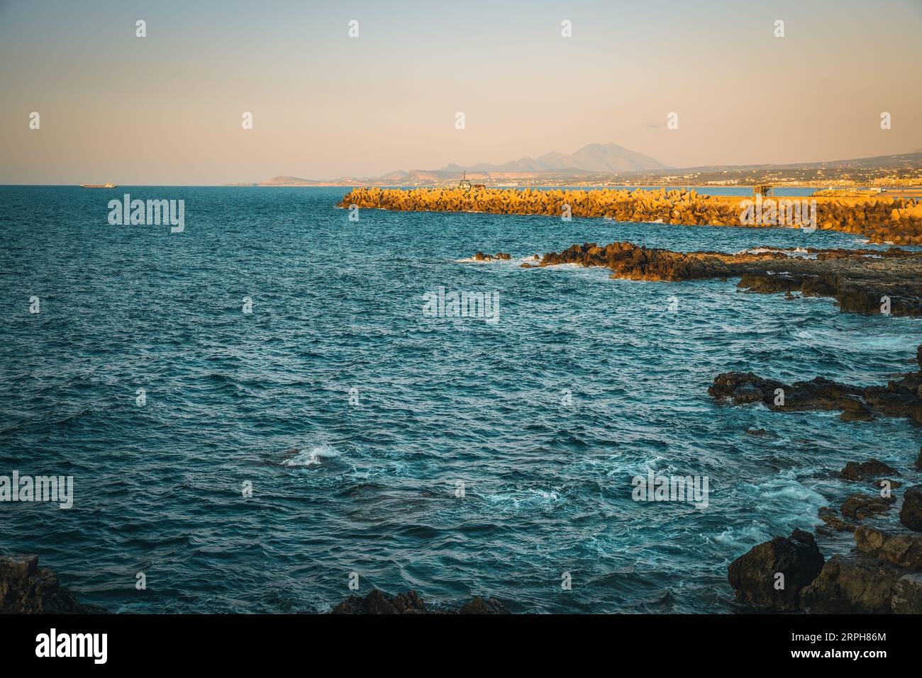 Experience the enchantment of twilight on a rocky beach in Rethymno. Against the backdrop of majestic mountains and with a ship gliding across the sea Stock Photo