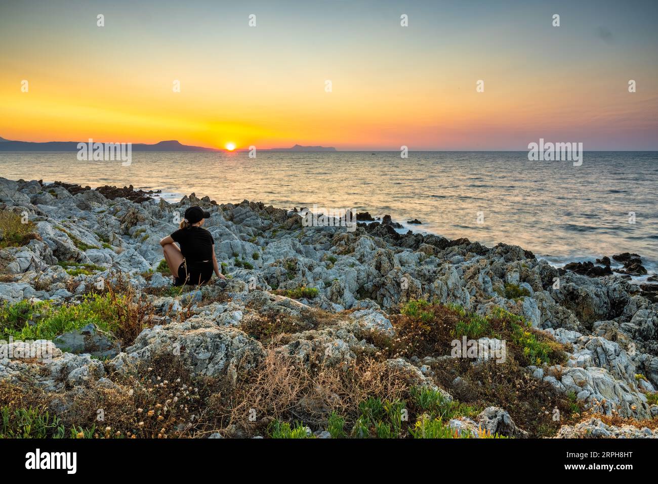 Capture the essence of quiet contemplation as a young girl sits upon the rocks, gazing at the sun's descent over Rethymno's rocky beach. The merging c Stock Photo