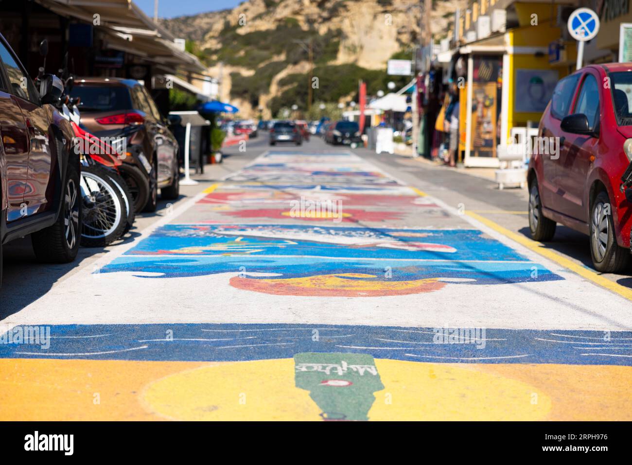 Bask in the vibrant spirit of Matala as you stroll through this sun-kissed street adorned with psychedelic hippie graphics. In the distance, the mount Stock Photo