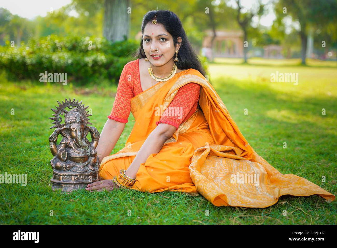 Happy young indian woman wearing saree sitting along with lord Ganesha idol outdoor at park celebrating Ganesh Chaturthi festival. Stock Photo
