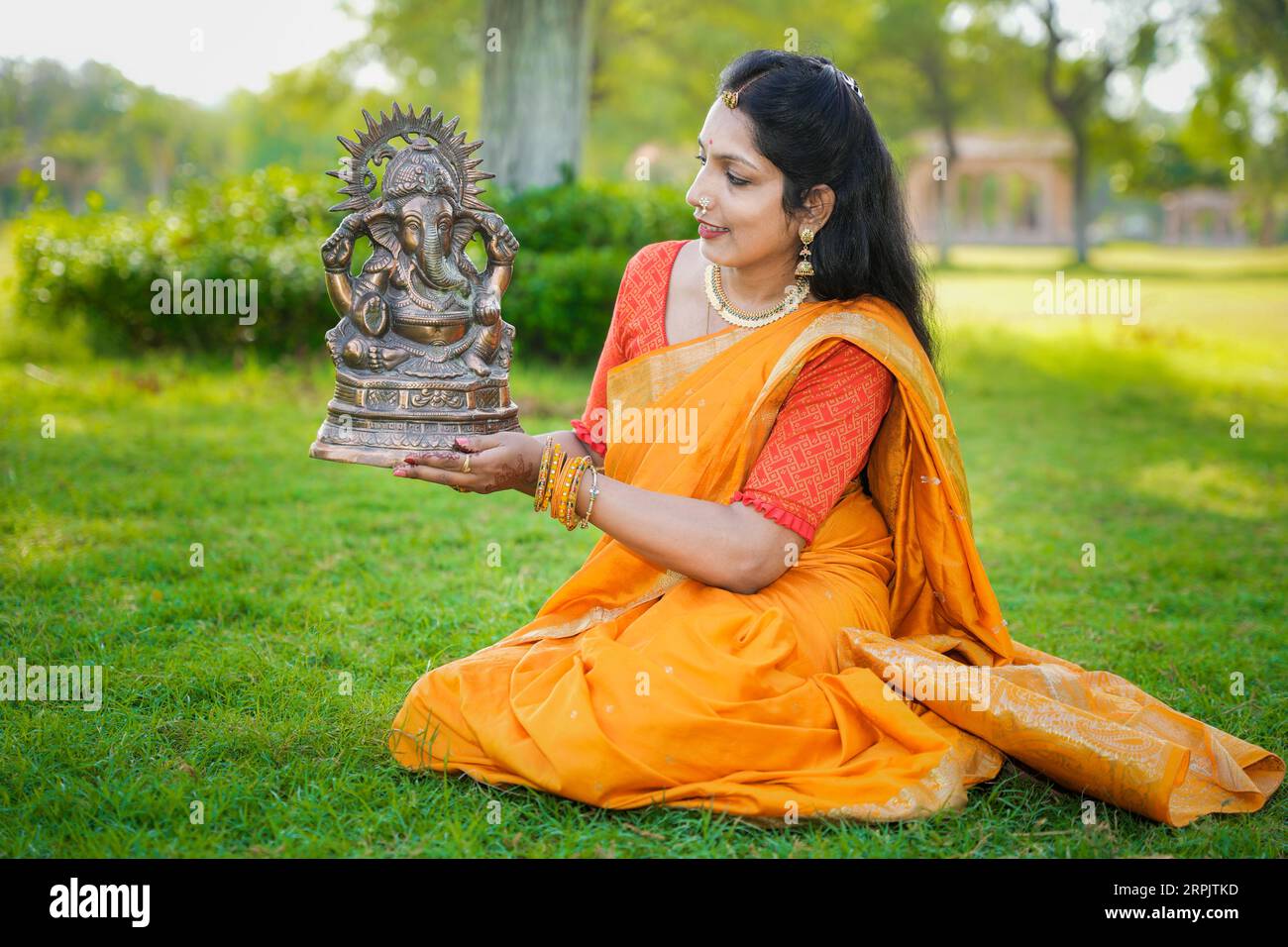 Happy young indian woman wearing saree sitting holding lord Ganesha idol outdoor at park celebrating Ganesh Chaturthi festival. Stock Photo