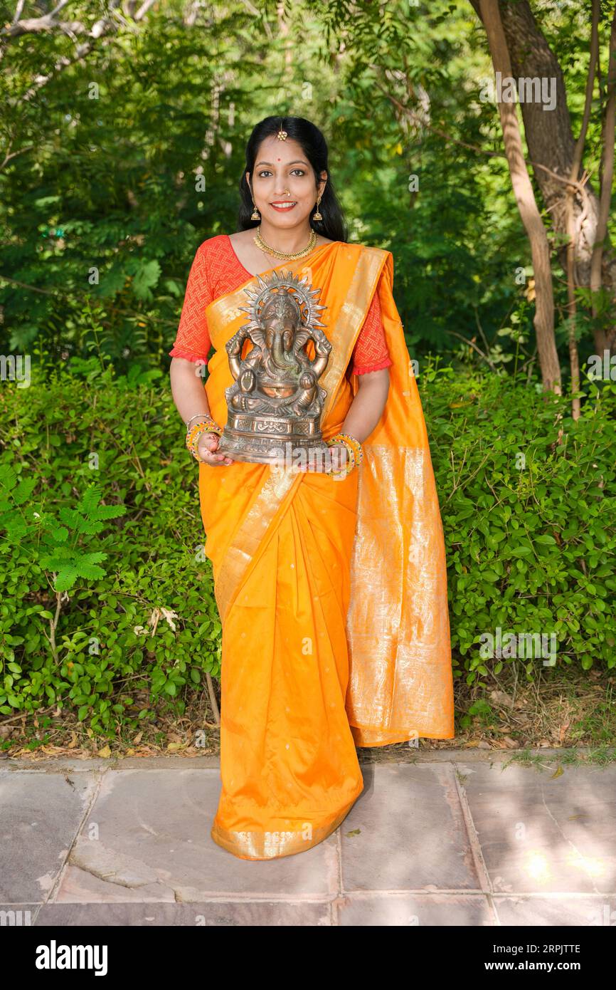 Happy young indian woman wearing saree holding lord Ganesha idol outdoor at park celebrating Ganesh Chaturthi festival. Stock Photo