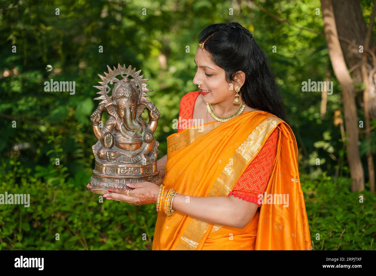 Happy young indian woman wearing saree holding lord Ganesha idol outdoor at park celebrating Ganesh Chaturthi festival. Stock Photo