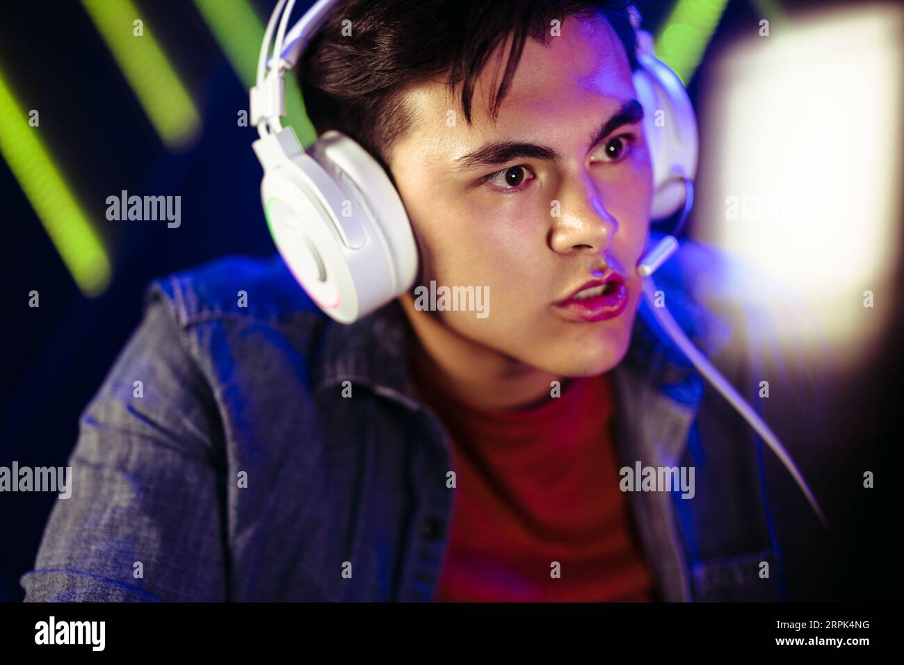 Young male gamer sitting with his eyes fixed on the computer screen in front of him. Wearing a headset, his expression is tense and his eyebrows are f Stock Photo