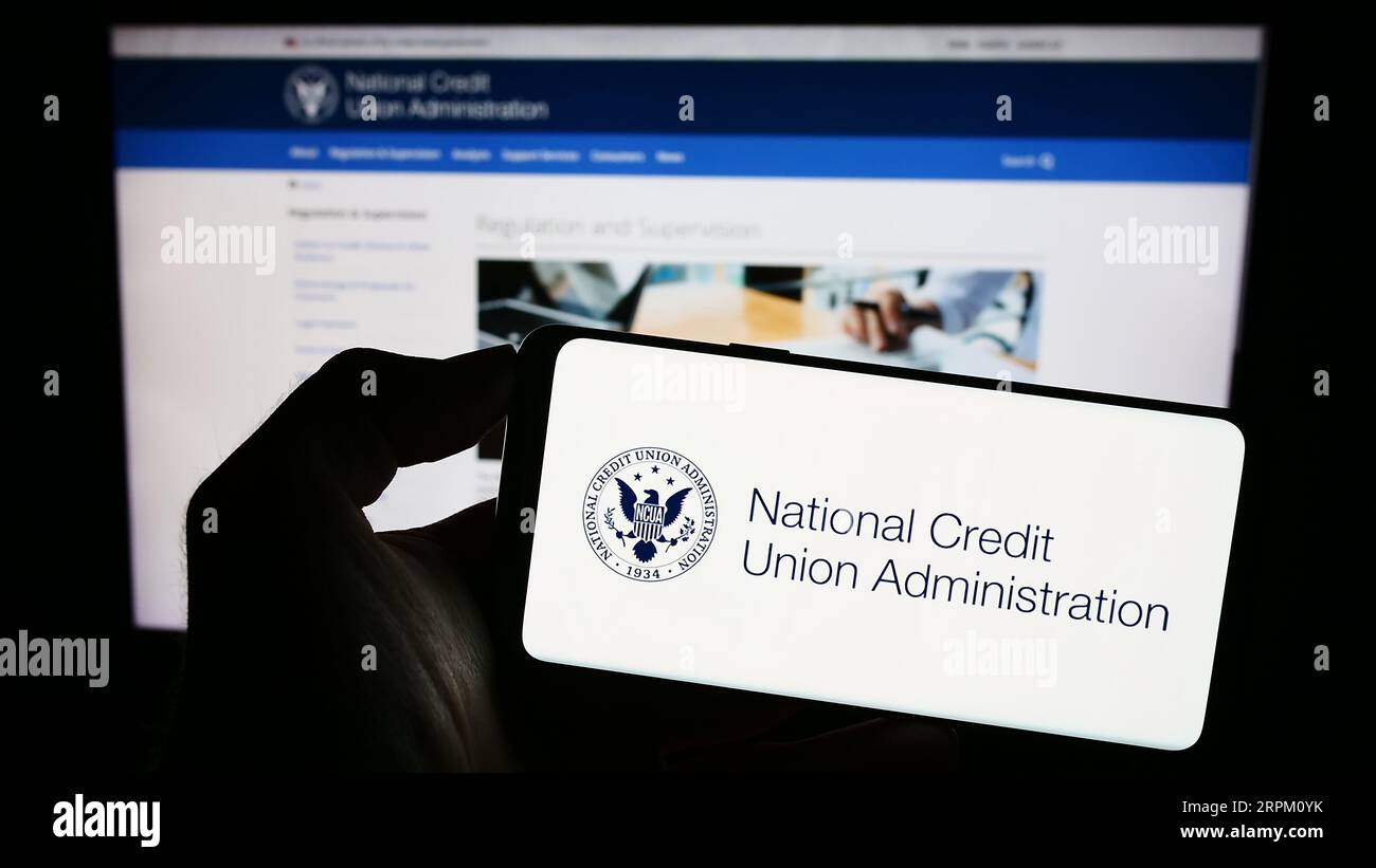 Person holding mobile phone with seal of National Credit Union Administration (NCUA) on screen in front of web page. Focus on phone display. Stock Photo