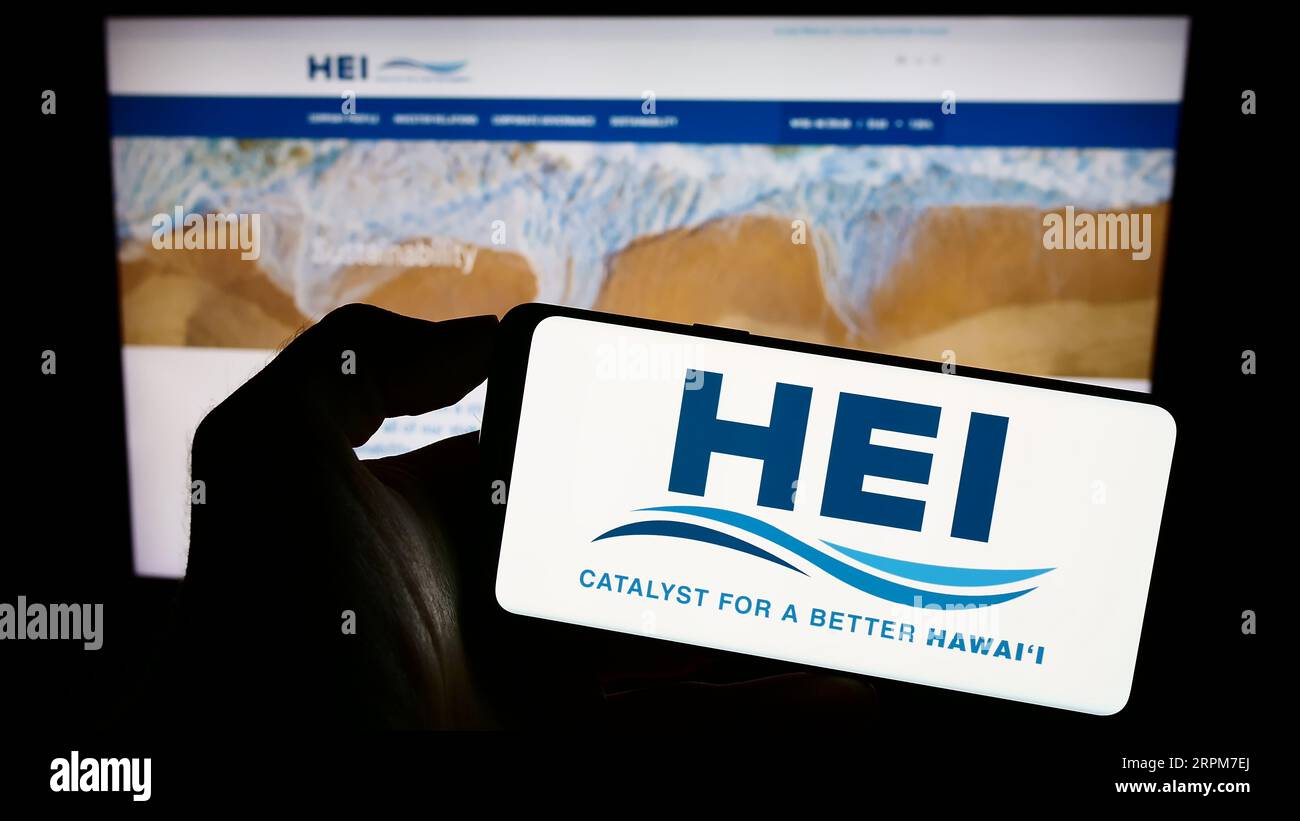 Person holding mobile phone with logo of company Hawaiian Electric Industries Inc. (HEI) on