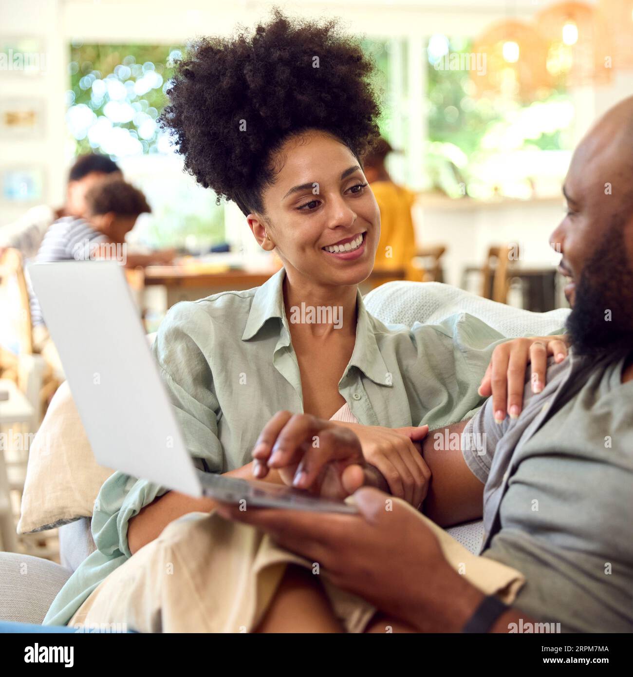 Couple At Home Using Laptop To Shop Or Book Holiday With Multi-Generation Family In Background Stock Photo