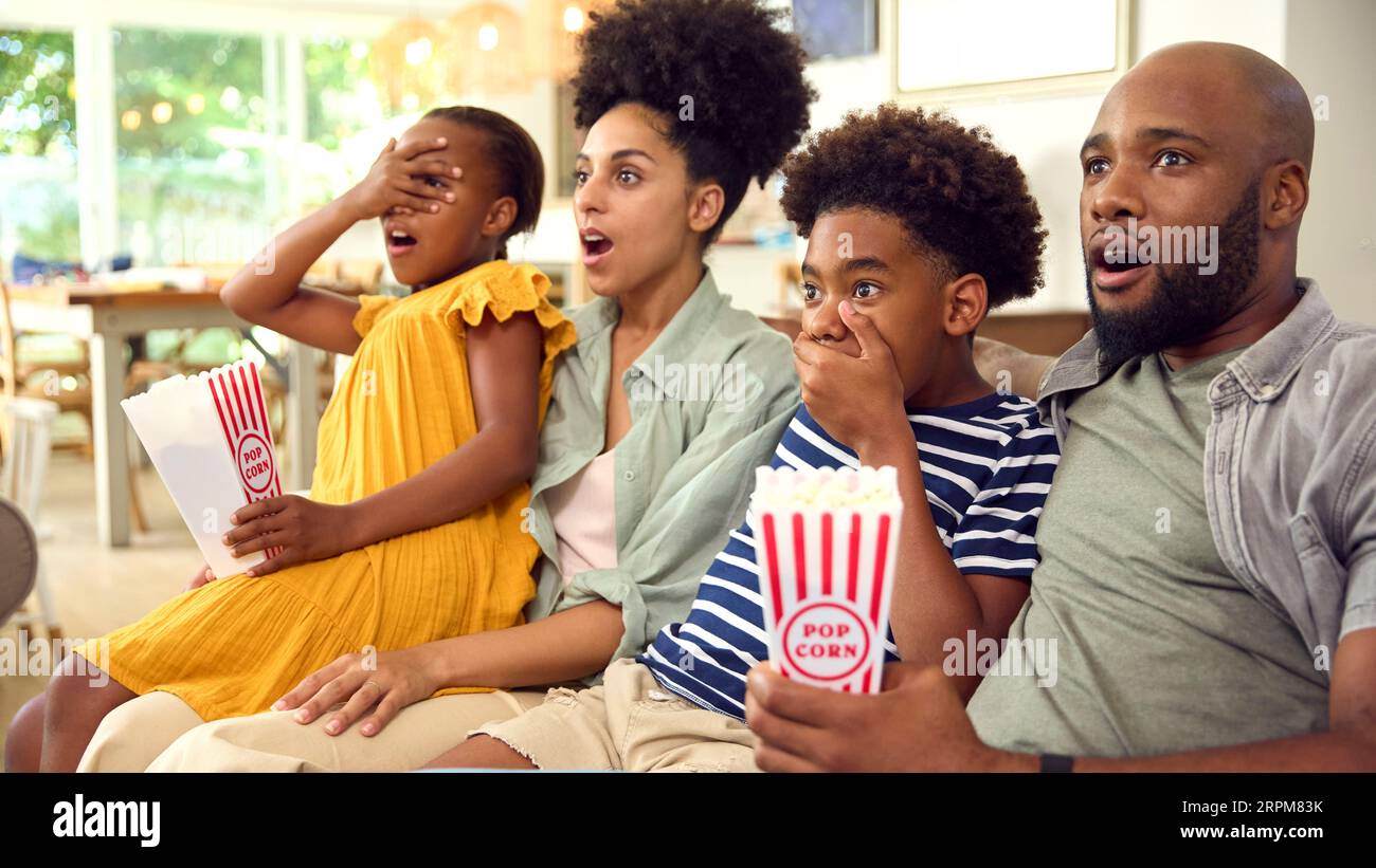 Frightened Family Eating Popcorn At Home Sitting On Sofa Streaming Horror Show Or Movie To TV Stock Photo