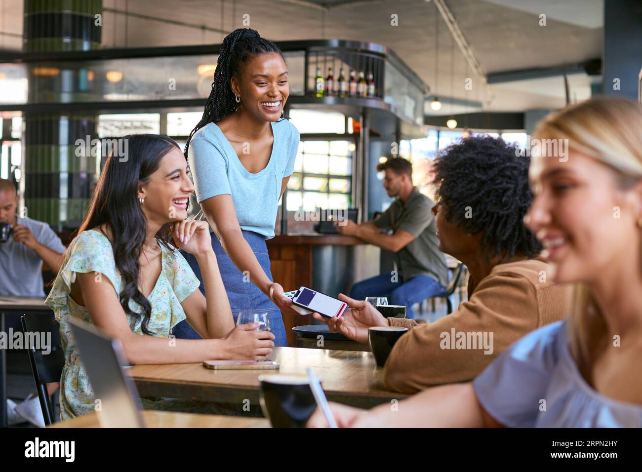 Couple In Coffee Shop Meeting With Man Paying Bill With Contactless Mobile Phone Payment Stock Photo