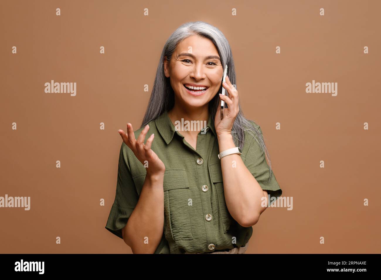 Cheerful happy mature Asian woman talking on smartphone isolated on brown background, 40s lady having pleasant phone conversation, chatting with friend Stock Photo