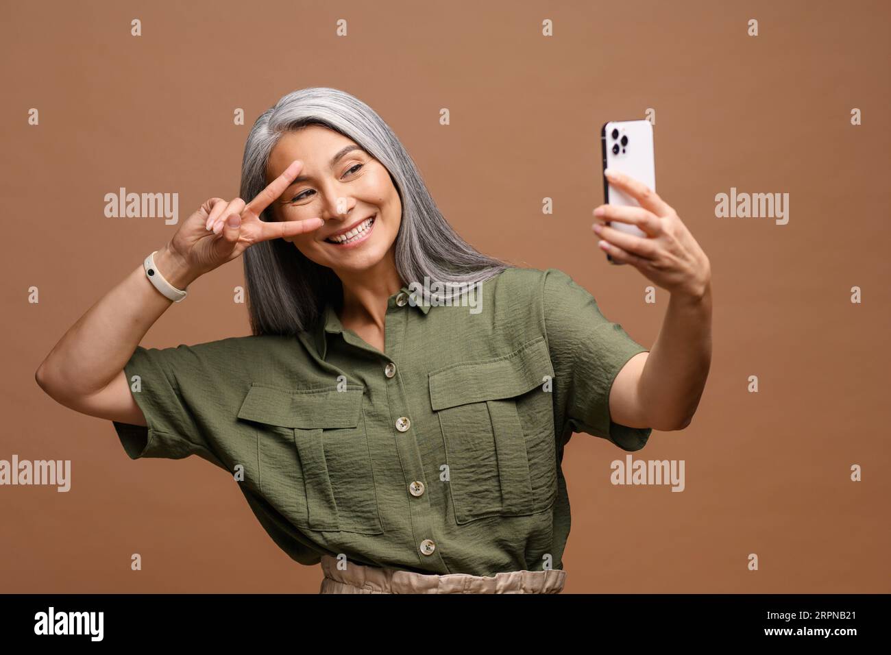 Mature Asian woman wearing casual green shirt with gray long hair standing isolated on brown background, taking selfie photo on smartphone looking camera laughing happy close-up Stock Photo