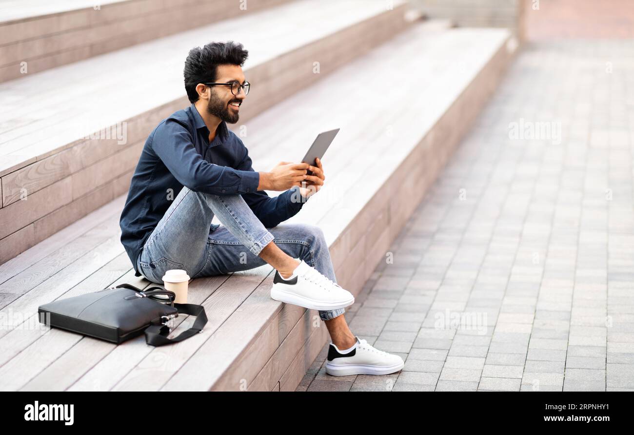 Relaxed indian guy chilling outdoor after work, using digital tablet Stock Photo