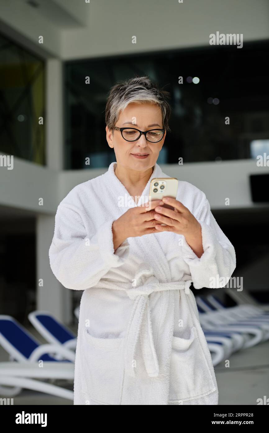 middle aged woman in white robe and glasses using smartphone inside of indoor spa center, retreat Stock Photo
